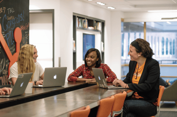 Three business women sitting at a table with their computers and laughing.